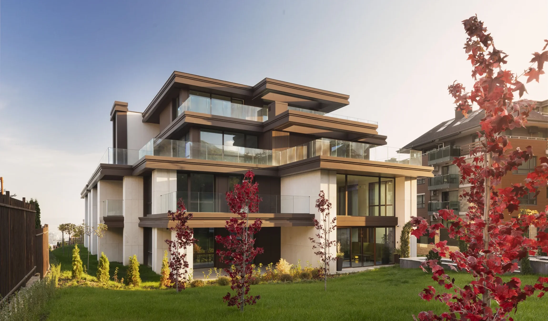 M Residence East наесен — нискоетажна жилищна сграда с широки дървени стрехи и стъклени парапети, червенолистно дърво пред нея и планински склон в дъното.