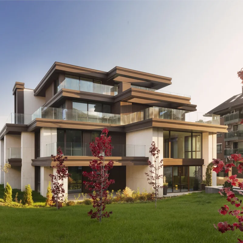 M Residence East наесен — нискоетажна жилищна сграда с широки дървени стрехи и стъклени парапети, червенолистно дърво пред нея и планински склон в дъното.