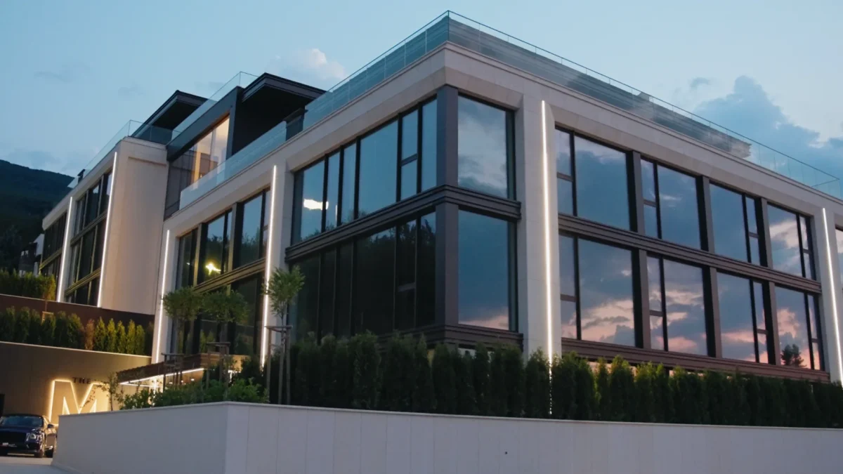 Жилищната сграда M Residence по здрач — стъклена фасада, която отразява залеза, светещ монограм „M“ на входа и паркирани автомобили пред нея.