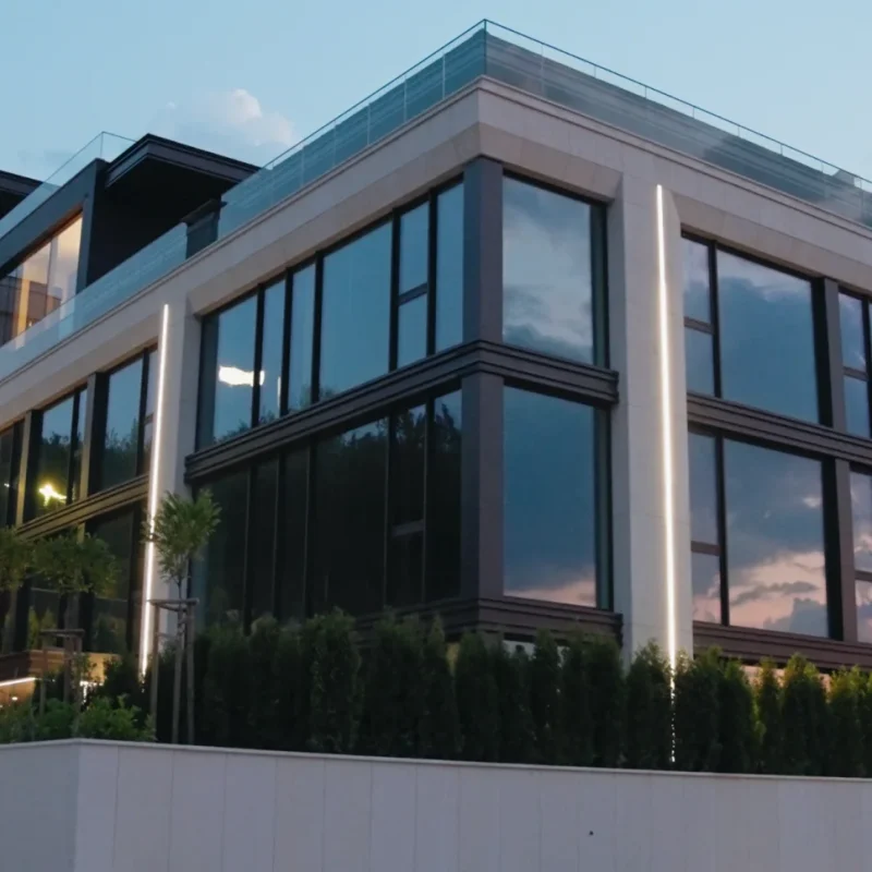 Жилищната сграда M Residence по здрач — стъклена фасада, която отразява залеза, светещ монограм „M“ на входа и паркирани автомобили пред нея.
