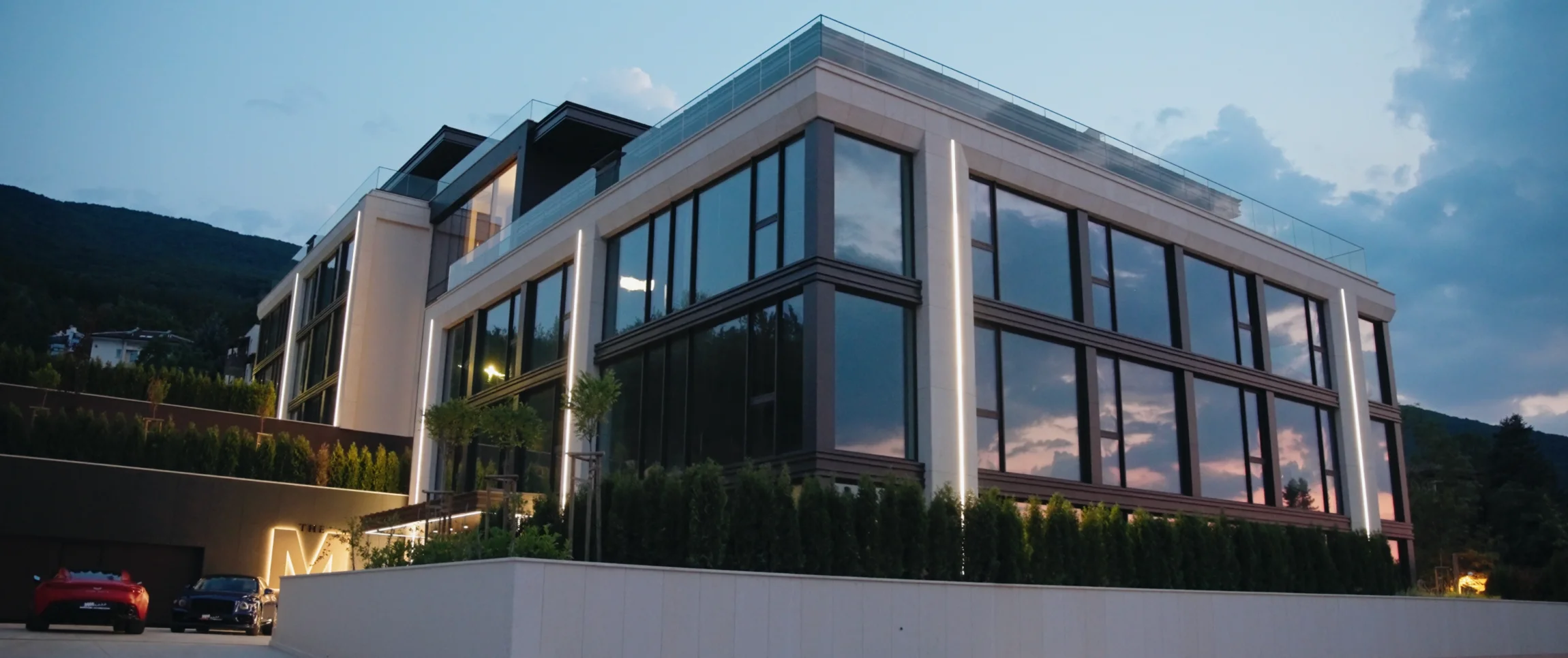 Жилищната сграда M Residence по здрач — стъклена фасада, която отразява залеза, светещ монограм „M“ на входа и паркирани автомобили пред нея.