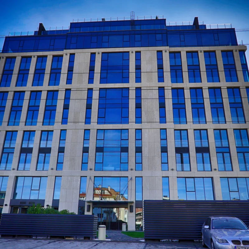 Сградата MM World отпред — каменни пилони и синьо остъкляване на осем етажа, вход в основата и автомобили пред нея.