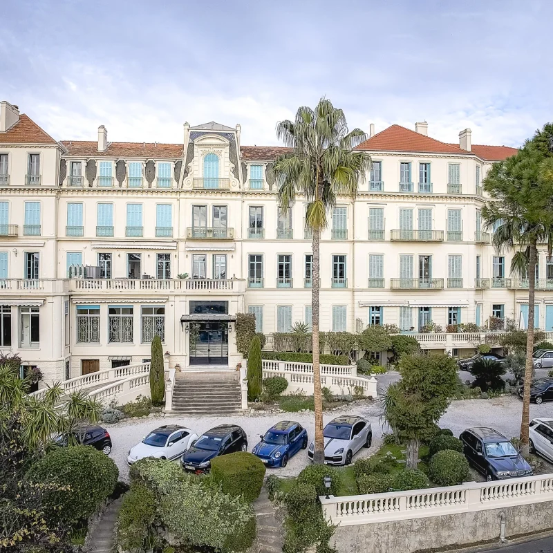 Palais Beauséjour в Кан — фасада в стил Бел епок със светли капаци и палми, стълбище към централния вход.